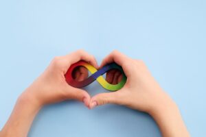 Can You Be A Little Bit Autistic? 19 Hands making a heart shape with a rainbow infinity sign inside the heart.