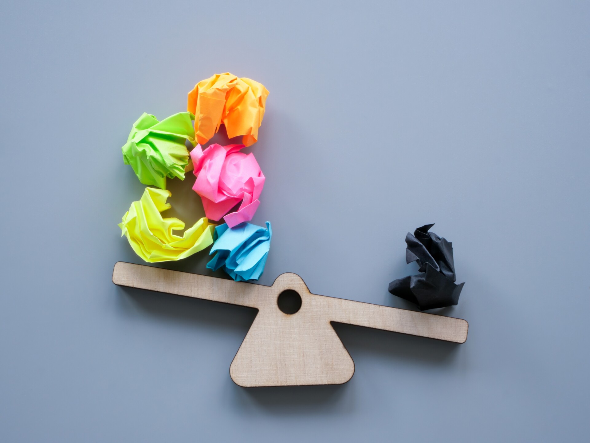 Negativity Bias A wooden seesaw with colorful paper balls on one side being weighed down by one black coloured ball of paper