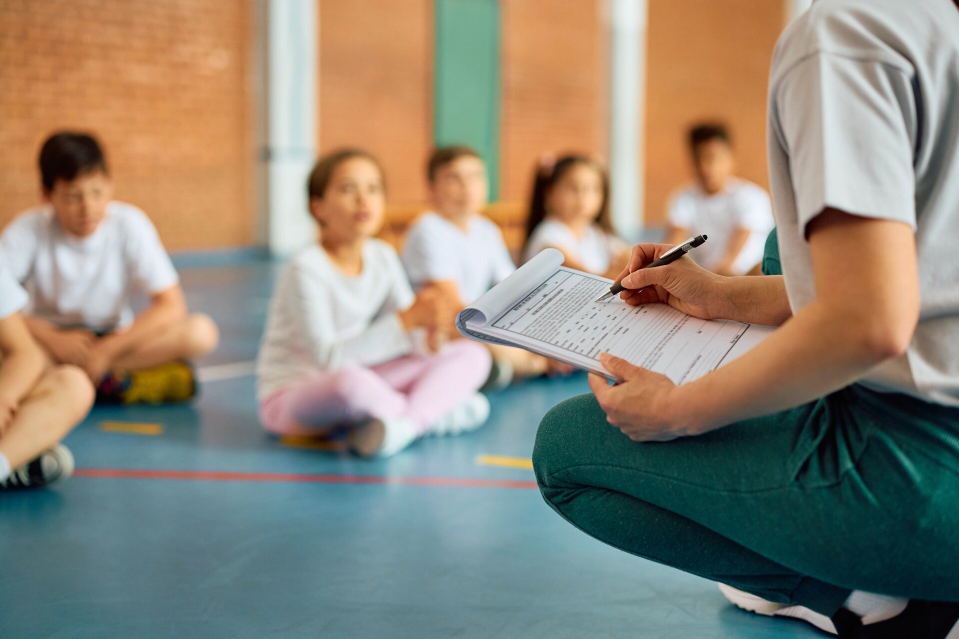 Physical Education Close up of a sports teacher writing on a clipboard with a gym full of PE students.